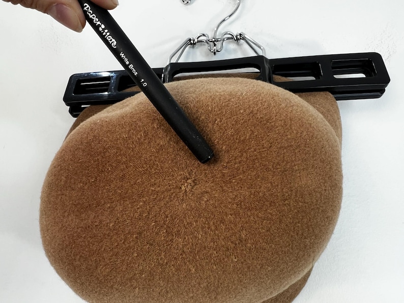 Pode incluir: Uma boina castanha pendurada num cabide preto. Uma caneta preta com o texto "Paper Mate Write Bros 1.0" aponta para a boina. A boina &eacute; feita de um material macio e felpudo.