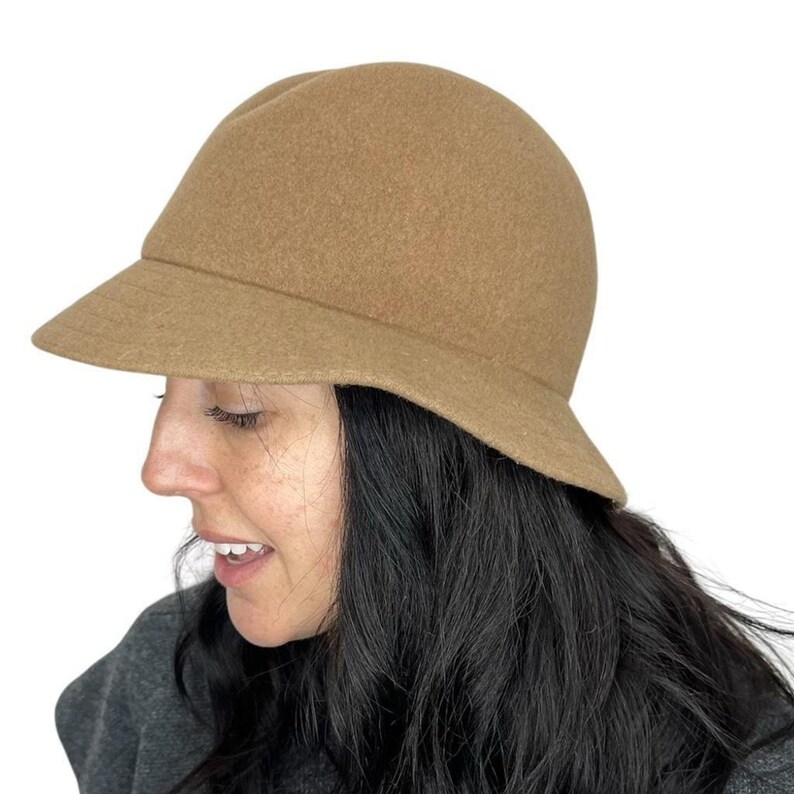 Pode incluir: Um chap&eacute;u cloche bege com uma aba pequena. O chap&eacute;u &eacute; feito de um material macio semelhante ao feltro. O chap&eacute;u &eacute; de cor neutra e pode ser usado com muitas roupas. O chap&eacute;u &eacute; um estilo cl&aacute;ssico.