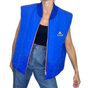 May include: A vibrant blue quilted vest with a ribbed collar and a full-length zipper. The vest features a white triangle logo with an upward arrow on the chest. The zipper has a coral-colored trim. The vest is sleeveless.