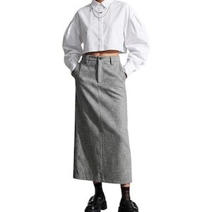 Peut inclure: Une chemise blanche courte à manches longues avec un col et une fermeture boutonnée, associée à une jupe longue grise en tweed à taille haute. La jupe présente une coupe droite, une couture verticale centrale et un bouton à la taille.