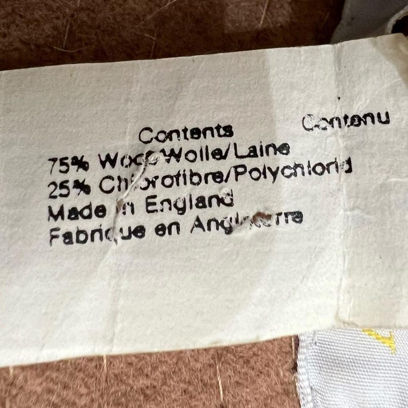 Pode incluir: Um close-up de uma etiqueta branca com texto preto. A etiqueta diz "Contents", "75% L&atilde;", "25% Clorofibra", "Made in England" e "Fabriqu&eacute; en Angleterre". A etiqueta est&aacute; ligeiramente rasgada.