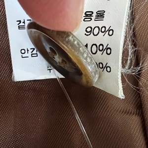 Puede incluir: Un bot&oacute;n redondo de color marr&oacute;n transl&uacute;cido con cuatro agujeros, sujeto por un hilo transparente a una etiqueta de cuidado de tela blanca. La etiqueta presenta texto impreso en negro, incluyendo la medida 96 cm y valores de 90% y 10%.