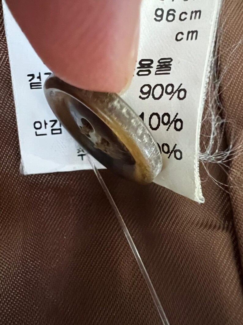 Puede incluir: Un bot&oacute;n redondo de color marr&oacute;n transl&uacute;cido con cuatro agujeros, sujeto por un hilo transparente a una etiqueta de cuidado de tela blanca. La etiqueta presenta texto impreso en negro, incluyendo la medida 96 cm y valores de 90% y 10%.