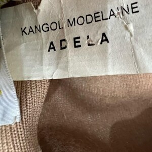 Pode incluir: Close-up de uma etiqueta com o texto "KANGOL MODELAINE ADELA" em preto. A etiqueta &eacute; esbranqui&ccedil;ada e ligeiramente enrugada. O fundo &eacute; um tecido de malha castanho claro.