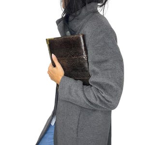 May include: A dark brown, textured snakeskin-patterned clutch bag with a metallic gold-tone frame edge, held against a charcoal gray wool-blend coat. The rectangular clutch features a distinct reptilian scale texture and a structured silhouette.
