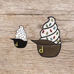 May include: Two embroidered patches of ice cream cones in a pirate hat. The larger patch is brown with a white ice cream cone with colorful sprinkles. The smaller patch is black with a white ice cream cone with colorful sprinkles.