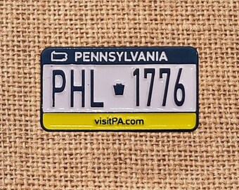 May include: A miniature Pennsylvania license plate pin with the text "PENNSYLVANIA", "PHL 1776", and "visitPA.com". The pin has a blue top, white center, and yellow bottom border, with black lettering.