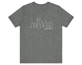 May include: A heather grey short-sleeve t-shirt featuring a white line art graphic of a city skyline integrated with a football trophy silhouette and the word CHAMPS printed in a clean, sans-serif font centred on the chest area of the garment.