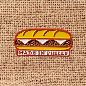 May include: A colorful pin shaped like a Philly cheesesteak sandwich. The pin features a yellow bun, red outline, and the text "MADE IN PHILLY" in white. The sandwich filling includes brown meat and white cheese.
