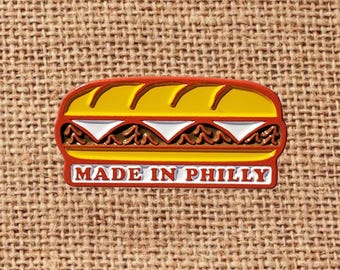 May include: A colorful pin shaped like a Philly cheesesteak sandwich. The pin features a yellow bun, red outline, and the text "MADE IN PHILLY" in white. The sandwich filling includes brown meat and white cheese.