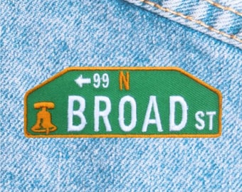 May include: A green and orange embroidered patch with the text "BROAD ST" and "99 N". The patch features a stylized image of a bell. The patch is on a light blue denim background.