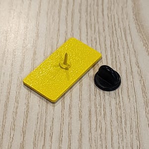 Puede incluir: Un pin de esmalte amarillo con un cierre de pin negro. El pin es rectangular y tiene una superficie lisa.