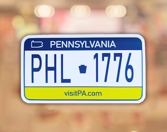 May include: A rectangular sticker resembling a Pennsylvania license plate. The sticker features a blue border, white background, and the text "PENNSYLVANIA" at the top. It displays "PHL - 1776" in blue and a yellow stripe with "visitPA.com".