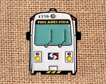 May include: A white and black enamel pin shaped like a Philadelphia train. The pin features the number "1776" and the word "PHILADELPHIA". The train has a teal window, yellow headlights, and a red, white, and blue logo.