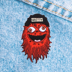 May include: A red embroidered patch featuring a cartoon monster with large orange eyes, a wide grin, and shaggy red fur. The monster wears a black hat with the word "GRITTY" in white letters. The patch is set against a blue denim background.