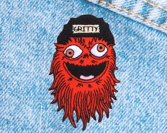 May include: A red embroidered patch featuring a cartoon monster with large orange eyes, a wide grin, and shaggy red fur. The monster wears a black hat with the word "GRITTY" in white letters. The patch is set against a blue denim background.