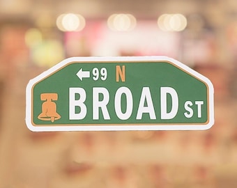 May include: A green and white Broad Street sign with the number 99 and an arrow pointing left. The sign has a bell graphic and is a street sign.