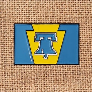 May include: A rectangular pin featuring a blue and yellow design with a stylized image of a bell. The pin has a black border and is set against a textured, tan background.