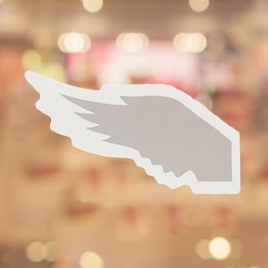 May include: A white and gray wing-shaped decal with a detailed feather design. The decal has a clean, modern aesthetic, suitable for various decorative applications. The background is blurred with warm tones.