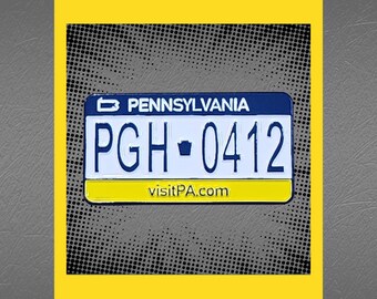 May include: A blue and yellow enamel pin featuring a Pennsylvania license plate with the text "PGH-0412" and the website "visitPA.com".