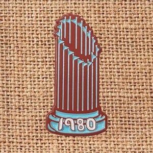 May include: A brown and blue enamel pin shaped like a trophy with the year "1980" on the base. The pin features vertical lines and small flags, set against a textured background.