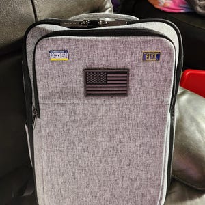 Puede incluir: Mochila gris con cremallera negra y correa negra. La mochila tiene un peque&ntilde;o parche de la bandera estadounidense y dos peque&ntilde;os parches azules y blancos que dicen "PGH-0412" y "You Got a Friend in PITT".