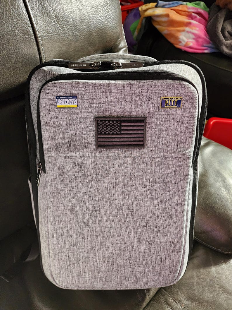 Puede incluir: Mochila gris con cremallera negra y correa negra. La mochila tiene un peque&ntilde;o parche de la bandera estadounidense y dos peque&ntilde;os parches azules y blancos que dicen "PGH-0412" y "You Got a Friend in PITT".