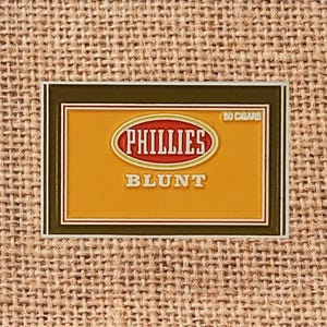 May include: A rectangular Phillies Blunt cigar box label with a brown border and a gold background. The Phillies logo is in red and white, with the word "BLUNT" in white below. The label reads "80 Cigars".