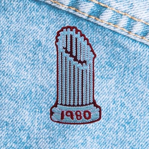 May include: Embroidered patch featuring a light blue and maroon design of a stylized monument. The patch has a maroon border and the year "1980" in red. The patch is on a light blue denim background.