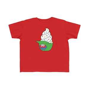 May include: A red t-shirt featuring a cartoon ice cream cone design. The ice cream is white with colorful sprinkles, and it's inside a green bowl with a blue monster face. The shirt is made of a soft material.