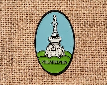 May include: Oval-shaped enamel pin featuring a white monument on a green base against a light blue sky. The pin has a black border and the word "PHILADELPHIA" in white text.