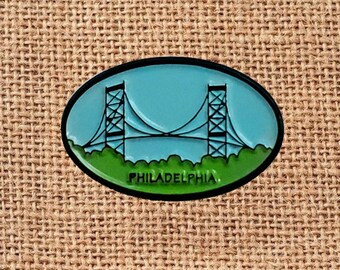 May include: An oval-shaped pin featuring a teal blue sky with a suspension bridge design. The bridge is black, and the bottom has a green grassy area. The word "PHILADELPHIA" is written in white at the bottom, with a black border.