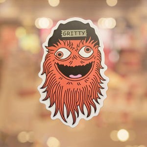 May include: A sticker featuring a cartoonish orange creature with a black hat that reads "GRITTY." The creature has large, expressive eyes, a wide grin, and a shaggy, orange beard. The sticker has a white border.