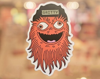 May include: A sticker featuring a cartoonish orange creature with a black hat that reads "GRITTY." The creature has large, expressive eyes, a wide grin, and a shaggy, orange beard. The sticker has a white border.