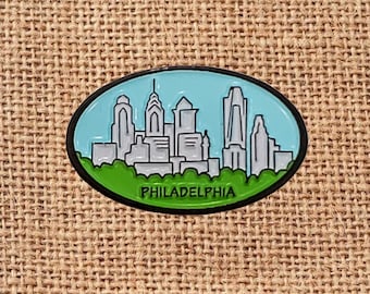 May include: Oval-shaped Philadelphia skyline pin with a black border. The pin features a light blue sky, green grass, and gray buildings. The word "PHILADELPHIA" is printed in black at the bottom.