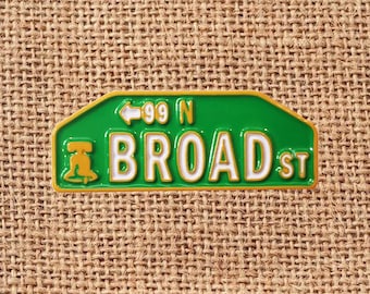 May include: A green and gold street sign pin with the text "BROAD ST" in white letters. The sign also features the number "99 N" and a bell symbol. The pin is set against a textured, tan background.