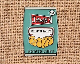 May include: A teal-colored pin shaped like a bag of potato chips. The pin features the text "JAWN'S" in red and white, "CRISP 'N TASTY" in a speech bubble, and "POTATO CHIPS" at the bottom. A small circle indicates a price of 25¢. The chips are yellow.