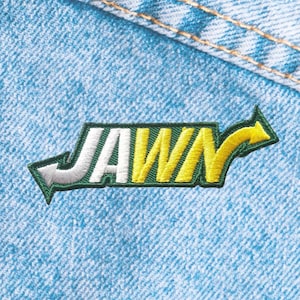 May include: Embroidered patch with the word "JAWN" in white and yellow, outlined in green, with arrows on each side. The patch is on a light blue denim background.
