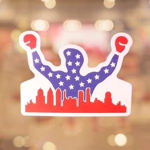 May include: A sticker featuring a silhouette of a muscular figure with arms raised, wearing red boxing gloves. The figure's body is filled with white stars on a blue background. A red skyline is at the bottom.