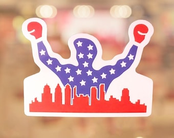 May include: A sticker featuring a silhouette of a muscular figure with arms raised, wearing red boxing gloves. The figure's body is filled with white stars on a blue background. A red skyline is at the bottom.