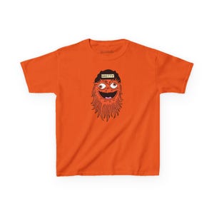 Op de afbeelding: Fel oranje t-shirt met een cartoon illustratie van een mascotte. De mascotte heeft oranje haar, grote ogen en een lachende mond. De zwarte hoed heeft de tekst "GRITTY".