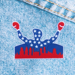 May include: A patriotic patch featuring a silhouette of a figure with raised arms, wearing boxing gloves. The figure is filled with white stars on a blue background. A red skyline and boxing gloves complete the design, set against a denim background.