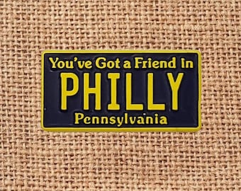 May include: A rectangular pin with a black background and yellow lettering. The text reads "You've Got a Friend in PHILLY Pennsylvania". The pin has a yellow border and is set against a textured, tan background.