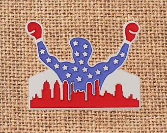 May include: A patriotic pin featuring a silhouette of a person with arms raised, wearing boxing gloves. The figure is blue with white stars, and the gloves are red. A red and white cityscape forms the base.