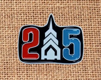 May include: A black and white enamel pin with the numbers "2" in red and "5" in blue. The numbers are outlined in white and black. The pin also features a white architectural design.