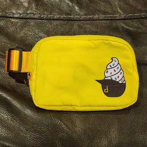 May include: A bright yellow fanny pack with a white and brown ice cream cone with sprinkles and a pirate hat embroidered on the front.
