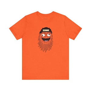 Könnte beinhalten: Orangefarbenes T-Shirt mit einer Cartoon-Illustration eines Maskottchens. Das Maskottchen hat orangefarbenes Fell, ein breites Grinsen und große Augen und trägt einen schwarzen Hut mit dem Namen "GRITTY". Das T-Shirt hat eine einfarbige orange Farbe.