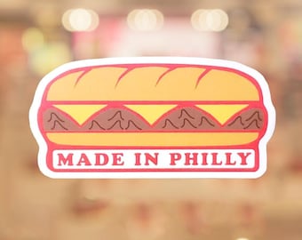 May include: A sticker featuring a cartoon illustration of a sandwich with a yellow bun, cheese, and meat. The sticker has a red border and the text "MADE IN PHILLY" in red letters.