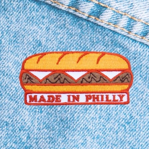 May include: Embroidered patch featuring a sandwich design with a yellow bun, brown filling, and white cheese. The patch has a red border and the text "MADE IN PHILLY" in white.