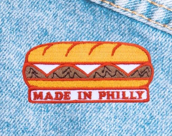 May include: Embroidered patch featuring a sandwich design with a yellow bun, brown filling, and white cheese. The patch has a red border and the text "MADE IN PHILLY" in white.